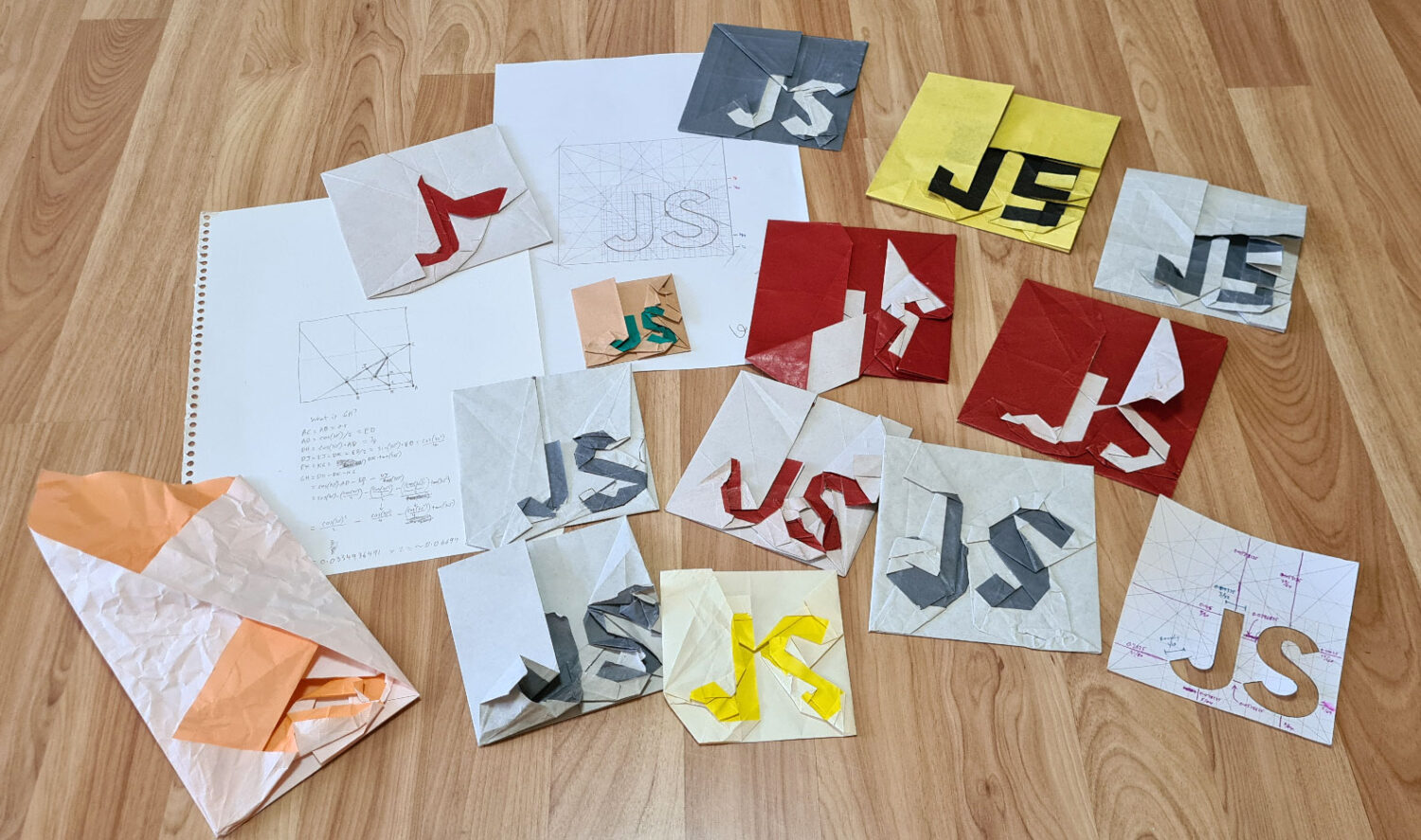 13 variations of origami JS logo attempts are scattered among printouts and sketches of shapes. Most of the models are rough and out-of-proportion. Some are only half folded.